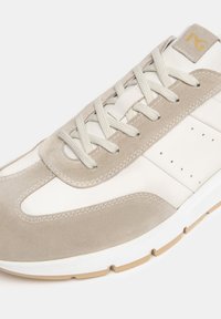 Zapatilla de ante beige y cuero blanco con tonos beige, diseño de cordones, detalles perforados y suela de goma en tono marrón claro.