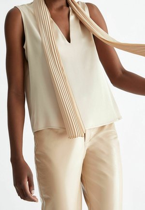 Kobieta w kremowej, bezrękawnikowej bluzce z dekoltem w serek, plisowanym beżowym szaliku i jasnobrązowych skórzanych spodniach na białym tle.