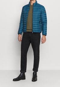Veste matelassée bleue avec un col haut, des zips, une poche d'accent, portée sur un pull vert olive, associée à un jean noir fuselé et des bottes.