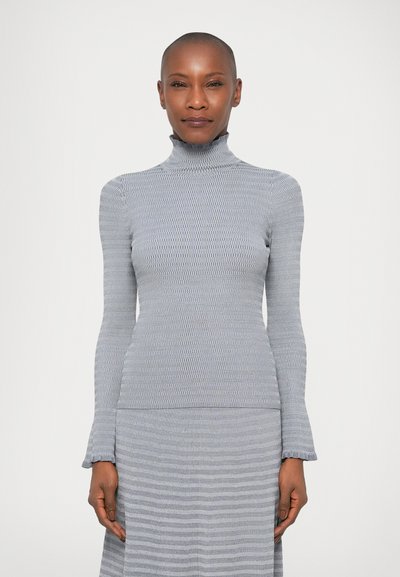 Femme aux cheveux très courts portant un haut gris à manches longues et col montant en côte ainsi qu'une jupe assortie, se tenant devant un fond blanc.