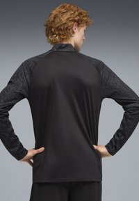 Camisa deportiva negra de manga larga con un torso liso y mangas texturizadas estampadas, con un corte relajado. Sin logotipos ni herrajes visibles.