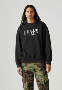 Felpa nera Levi's con logo bianco, caratterizzata da un collo rotondo e polsini a coste, abbinata a pantaloni cargo camuffati.