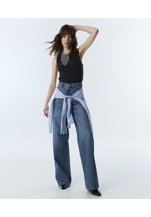 Jeune femme aux cheveux bruns lâchés porte un débardeur noir, un jean bleu à jambes larges, une chemise rayée nouée à la taille et des chaussures noires pointues.