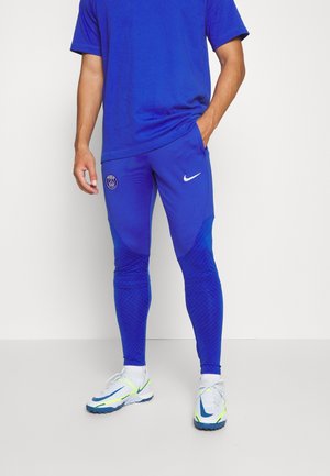 Mand iført blå sportbukser med Paris Saint-Germain og Nike logoer, hvide sokker og hvide løbesko med blå og grønne detaljer.