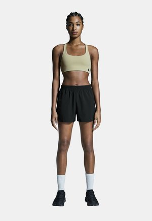Zwarte sportshorts en een lichtbruine sportbeha, gecombineerd met kniehoge witte sokken en zwarte sneakers, gedragen door een persoon die rechtop staat.