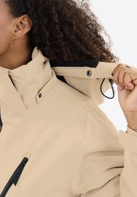 Beige waterdichte jas met hoge kraag, verstelbare capuchon, ritszakken en gestructureerde stof. Bevat drukknoop- en trekkoorddetails.