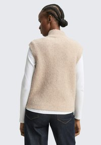 Beige gebreide vest met een hoge kraag, voorzien van een gestructureerd patroon. Gekozen over een wit langshirt, in combinatie met donkere jeans.