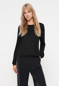 Schwarzes Langarmoberteil mit einem strukturierten Leopardenmuster, rundem Ausschnitt und lässiger Passform. Kombiniert mit glänzenden schwarzen Hosen.