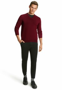 Burgunderfarbener Strickpullover mit V-Ausschnitt, kombiniert mit schwarzen Slim-Fit-Hosen und schwarzen Sneakers mit grauen Socken. Einfaches, zeitgemäßes Design.
