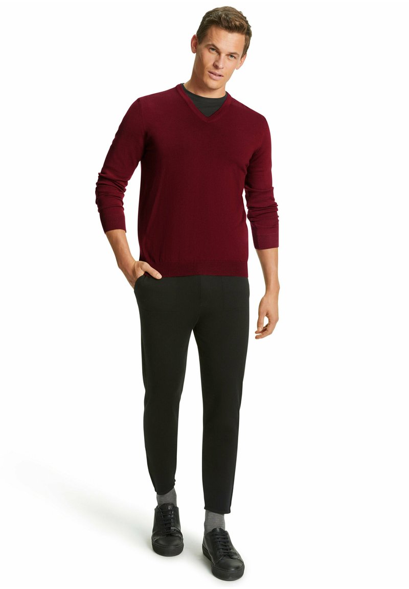 Burgunderfarbener Strickpullover mit V-Ausschnitt, kombiniert mit schwarzen Slim-Fit-Hosen und schwarzen Sneakers mit grauen Socken. Einfaches, zeitgemäßes Design.