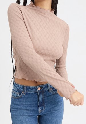 Kobieta ubrana w beżowy, teksturowany crop top z długim rękawem i falistymi krawędziami oraz niebieskie dżinsy z wysokim stanem, poprawia rękaw prawą ręką.