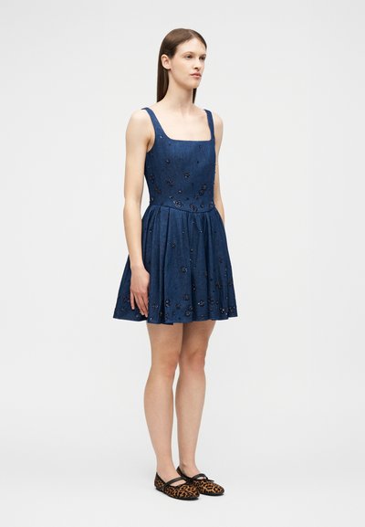 Γυναίκα που στέκεται με αμάνικο μπλε φόρεμα με διακοσμητικά λουλουδιών και επίπεδα παπούτσια με λεοπάρ μοτίβο, μπροστά σε απλό φόντο.