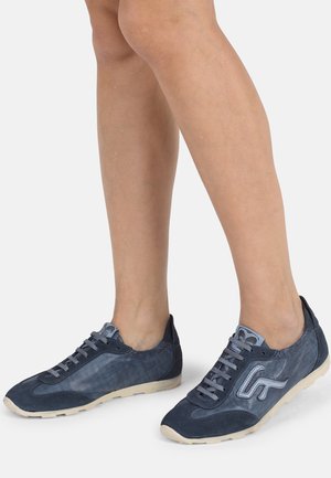 Nohy v tmavě modrých sportovních teniskách se světle béžovými podešvemi, zobrazené na jednobarevném světlém pozadí.