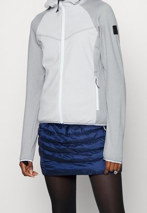 Frau trägt eine hellgraue Jacke mit Reißverschluss und Daumenlöchern sowie einen gesteppelten marineblauen Minirock über schwarzen Strumpfhosen.