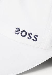 Fehér sapka ívelt karimával, szövet textúrával és feltűnő sötétkék "BOSS" logóval az elülső részen. Oldalt látható varrás részletek.