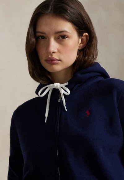 Νεαρή γυναίκα με κοντά καστανά μαλλιά που φοράει μπλε σκούρο φούτερ με φερμουάρ, λευκά κορδόνια και μικρό κόκκινο κεντητό λογότυπο στο στήθος.