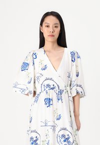 Weißes Kleid mit blauen Blumen- und geometrischen Mustern. Mit kurzen Puffärmeln, tiefem V-Ausschnitt und gerafftem Bund. Weicher Stoff.