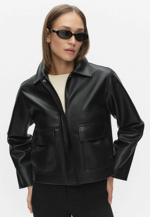 Femme portant une veste en similicuir noir avec de grandes poches avant et des lunettes de soleil ovales noires, posant les mains sur les hanches.