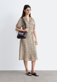 Woman standing in a beige floral dress, wearing sunglasses, black sandals, and carrying a dark brown crossbody bag against a plain background.