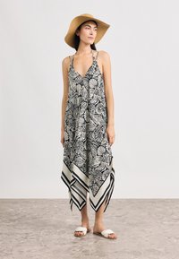 Μακρύ φόρεμα σε ασπρόμαυρο με floral σχέδιο, V-λαιμόκοψη, τιράντες halter, ασύμμετρο τελείωμα και εκρού λεπτομέρειες. Συνδυάζεται με λευκά σανδάλια και καφέ καπέλο.