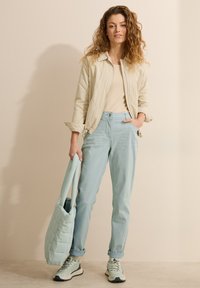 Chaqueta acolchada color beige claro, blusa cremosa, pantalones suaves color azul claro con bolsillos y bolso de mano teal a juego. Zapatillas con acentos oscuros.