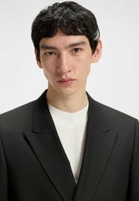 Schwarzer, maßgeschneiderter Blazer mit scharfen Revers, kombiniert mit einem schlichten weißen Hochkragenhemd. Glatte Textur mit strukturiertem Sitz.