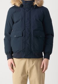 Marineblauwe pufferjack met faux fur trim aan de capuchon, twee klepzakken, elastische manchetten en ribgebreide onderkant. Glad stof en stevige ritsen.