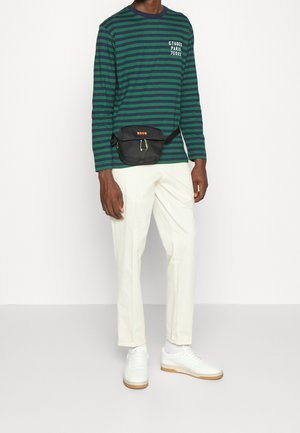 Camicia a maniche lunghe a righe verdi e blu navy con grafica bianca, pantaloni bianchi e sneakers bianche. Borsa a tracolla nera con logo arancione brillante.
