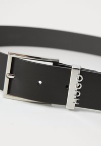 Ceinture en cuir noir avec une boucle rectangulaire en argent. Présente un accent logo et plusieurs trous pour l'ajustement de taille. Texture lisse.