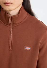 Smeđa dukserica sa zatvaračem koja ima visoki ovratnik, meku teksturu i mali "Dickies" logo pečat s lijeve strane.