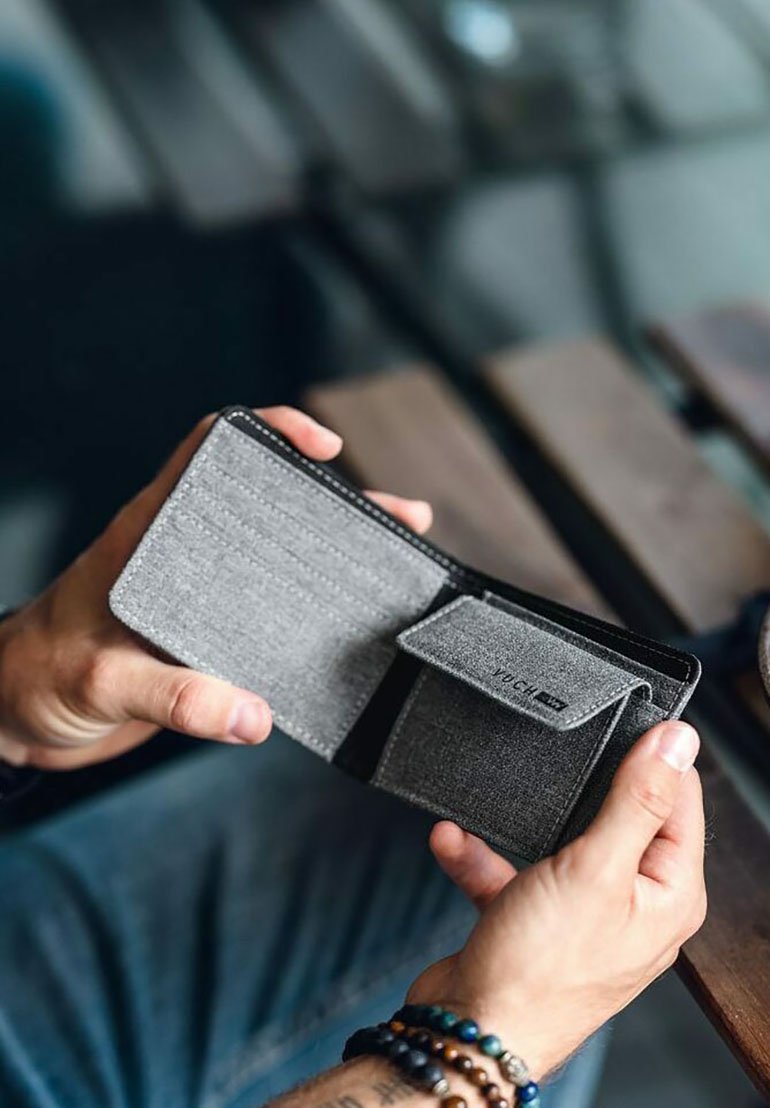 Šedá tkaninová peněženka otevřená v rukou, obsahující přihrádky na karty a kapsu s klopou. Hladká textura s kontrastními černými okraji a švy.