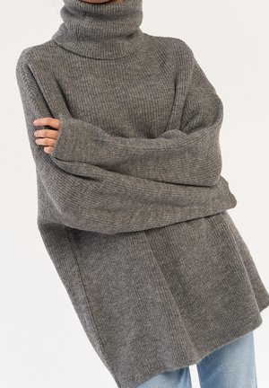 Persoon met een oversized grijze coltrui met lange mouwen, armen gekruist, staand tegen een effen lichte achtergrond.