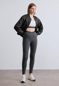 Μαύρο bomber μπουφάν πάνω από λευκό crop top, γκρι ψηλόμεσα κολάν με λογότυπο στη μέση και λευκά αθλητικά παπούτσια.