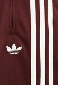 Μαρονοκό ύφασμα με λευκό λογότυπο Adidas trefoil και τρεις κάθετες λευκές ρίγες, με κάθετη τσέπη με φερμουάρ.