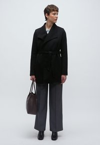Μαύρο παλτό με ζώνη και γιακά σαάλι, φορεμένο πάνω από ριγέ τοπ, συνδυασμένο με γκρι παντελόνι φαρδιάς γραμμής και καφέ πλεκτή τσάντα.