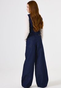 Denim wijde broek in donkerblauw, met een hoge taille en een ruime pasvorm. Achterzakken zichtbaar, aangevuld met een aansluitend vest erboven.