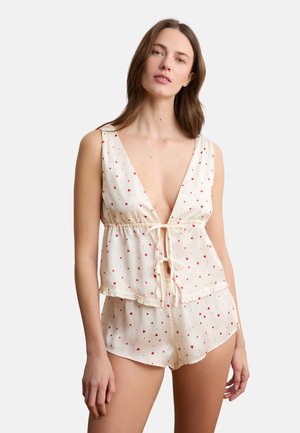 Femme portant un ensemble de pyjama couleur crème avec des imprimés de cœurs rouges, composé d'un haut sans manches à col en V profond noué à l'avant et d'un short assorti.