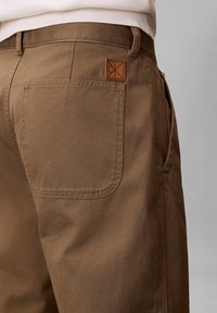 Close-up van de achterzak van een bruine broek met een leren label en zichtbare stiksels, gedragen met een wit shirt.