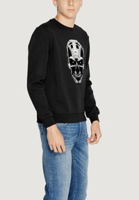 Sudadera negra de algodón con un diseño gráfico de calavera que presenta acentos metálicos y ojos verdes; puños y dobladillo ajustados, estilo casual.
