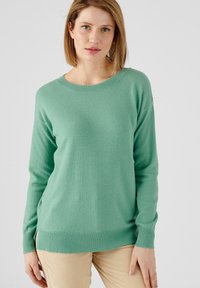 Suéter de punto verde claro con cuello redondo, mangas largas y dobladillo acanalado. Textura suave con un ajuste relajado, combinado con pantalones de pana beige.