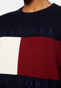 Pull marin arborant des blocs horizontaux de rouge et de blanc, texture côtelée, et broderie "TOMMY HILFIGER NEW YORK" en bas.