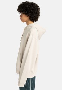 Crème-kleurige hoodie met ribgebreide boorden en zoom, een voorzak, een capuchon met trekkoord en een losse pasvorm, afgebeeld vanuit de zijkant.