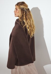 Kohvakarva pehme kangast ümbris, millel on lihtne disain, lahtine sobivus ja ümarad servad. Juuksed on stiliseeritud lahtiste lokkidega.