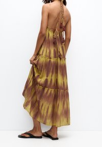 Robe longue à motif tie-dye avec des nuances de jaune et de marron, tissu léger, col halter avec liens tressés et accents de pompons.