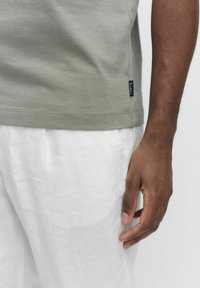 Olivgrön bomullströja med sidoflisksmärke, i kombination med vita linbyxor. Närbild visar texturerad tygkvalitet och avslappnad passform.