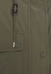 Tkanina w oliwkowym kolorze z przeszytymi szwami, klapka kieszeni zabezpieczona ciemnym zatrzaskiem oraz sznurek ze metalowym końcem i skórzaną pętelką.