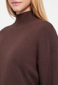 Brązowy sweter z wysokim prążkowanym kołnierzem, miękką teksturą i luźnym krojem, z szerokimi rękawami i bezszwową konstrukcją.