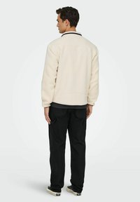 Mann steht mit dem Rücken zur Kamera, trägt eine cremefarbene Fleecejacke, schwarze Hosen mit Seitentaschen und schwarze Low-Top-Sneaker.