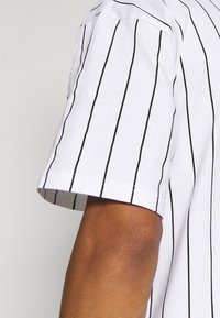 Bílá košile s krátkým rukávem a vertikálními černými pruhy. Má zaoblený výstřih a kapsu na hrudi na levé straně. Měkčená bavlněná tkanina.