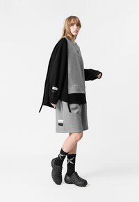 Sudadera gris de capa debajo de una sudadera negra de gran tamaño, combinada con pantalones cortos grises y zapatos de tela negra sin cordones. Calcetines negros con estampado blanco.